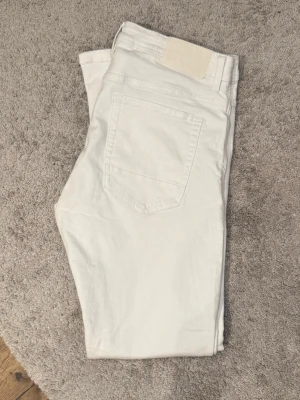 Vita raka slimfit jeans från smog demin - Säljer ett par vita jeans med rak slimfit passform och klassisk femficksdesign. Jeansen har silverfärgade nitar och knapp, samt en vit patch bak i midjan. Perfekta för en clean och stilren look.