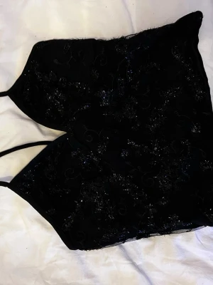 Svart glittrig korsettopp med spets - Snygg svart korsettopp med tunna axelband och glittriga detaljer. Toppen har ett elegant spetsmönster och figurnära passform som ger en trendig look. Perfekt att styla med jeans eller kjol för en cool outfit.