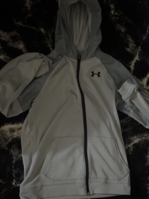 Ljusgrå hoodie från Under Armour - Snygg ljusgrå hoodie från Under Armour med dragkedja framtill och svart logga på bröstet. Hoodien har huva, fickor framtill och är tillverkad i ett mjukt, sportigt material. Perfekt för dig som gillar en clean och sportig stil.