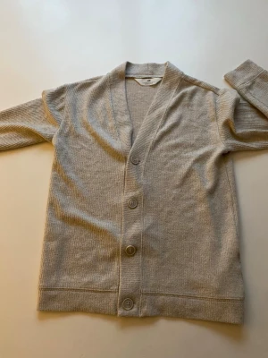 Beige ribbad kofta med knappar - Snygg beige kofta med ribbad struktur och tre stora knappar framtill. Koftan har V-ringning och långa ärmar, perfekt för lager på lager. Tillverkad i mjukt material som känns skönt mot huden.