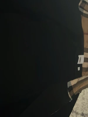 Svart hoodie från Burberry - Snygg svart hoodie från Burberry med dragkedja och den klassiska beige-rutiga huvan. Hoodien har långa ärmar och ribbade muddar. Perfekt för dig som vill ha en stilren look med en touch av lyx. Materialet känns mjukt och bekvämt.