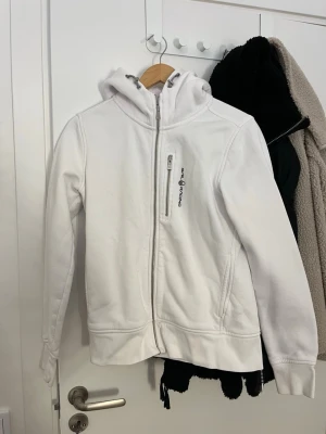 Vit hoodie från Peak Performance - Snygg vit hoodie från Peak Performance med dragkedja framtill och en bröstficka med logga. Hoodien har justerbar huva med snören och två sidofickor. Perfekt för en avslappnad och sportig stil.