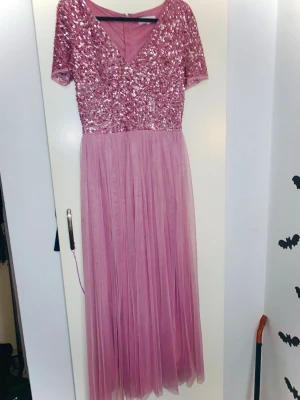 Rosa långklänning med paljetter - Långklänning i rosa med glittrig paljettöverdel och v-ringning. Kjolen är gjord i flera lager tunn tyll som ger ett svepande fall. Klänningen har korta ärmar och dragkedja i ryggen. Perfekt för speciella tillfällen när du vill sticka ut. Nypris ca 600kr