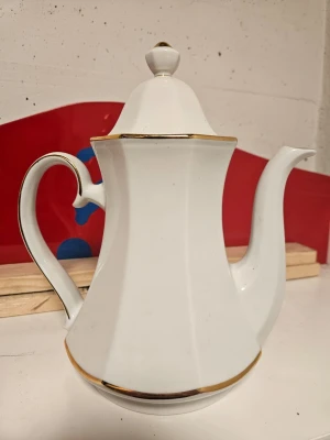 Vit porslinskanna med gulddetaljer - Elegant vit porslinskanna med lock och guldfärgade detaljer längs kanterna. Kannan har en klassisk, svängd form och ett dekorativt handtag. Perfekt för servering av kaffe eller te och ger en lyxig känsla till dukningen.