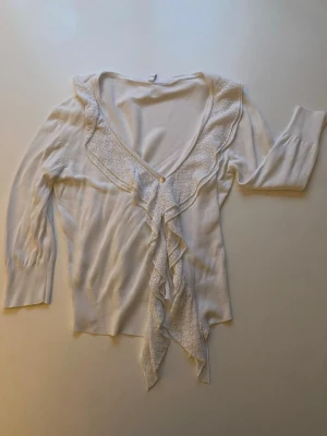 Vit volangkofta med spetsdetaljer - Söt vit kofta med djup V-ringning, volangkant och spetsdetaljer längs kragen. Koftan har långa ärmar, ribbade muddar och knytband framtill. Perfekt för en romantisk och feminin stil.