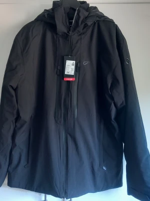 Svart vindjacka med huva från Five seasons - Stilren svart vindjacka med huva och hel dragkedja framtill. Jackan har två sidofickor med dragkedja och en bröstficka. Tillverkad i ett slitstarkt, vattenavvisande material och har justerbara ärmslut. Perfekt för blåsiga dagar.