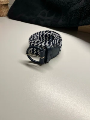 Blåvitt flätat bälte med läderdetaljer - Snyggt flätat bälte i blått och vitt med läderdetaljer vid spännet. Bältet har ett randigt mönster och ett klassiskt metallspänne. Perfekt accessoar för att lyfta din outfit.