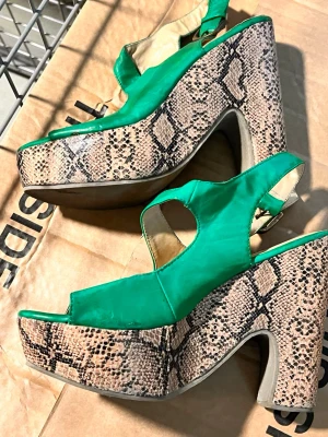 Gröna platåsandaler med ormmönster - Unika gröna sandaler med bred platå och hög klack i ormmönstrad design. Skorna har öppen tå, justerbart spänne vid ankeln och coola utskärningar på sidorna. Materialet ser ut att vara konstläder och textil. Perfekta för dig som vill sticka ut.