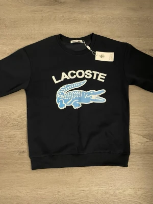Svart sweatshirt från Lacoste - Cool svart sweatshirt från Lacoste med stort blått och vitt krokodilmotiv och logga på bröstet. Klassisk rund halsringning och ribbade muddar. Perfekt för en avslappnad och trendig look.