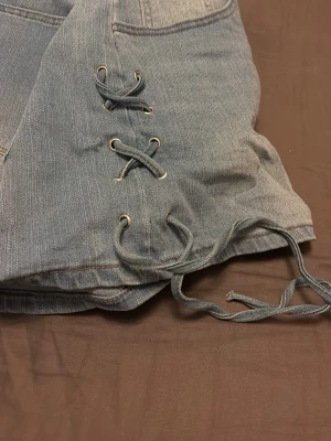 Blå jeansshorts med snörning - Snygga blå jeansshorts med snörning i sidorna och klassiska bakfickor. Shortsen har en avslappnad passform och är perfekta för varma dagar. Tillverkade i slitstarkt denimtyg med coola detaljer som ger en trendig look.