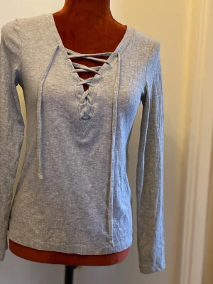 Ljusgrå långärmad topp med snörning XS - Trendig ljusgrå långärmad topp med ribbad struktur och snygg snörning framtill i urringningen. Perfekt för dig som gillar en avslappnad men ändå stilren look. Tillverkad i mjuk bomullsmix som känns skön mot huden.