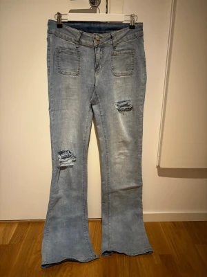 Ljusblå bootcut jeans med slitningar - Säljer ett par ljusblå bootcut jeans med slitningar på låret och knät. Jeansen har hög midja, två framfickor och två bakfickor med knappdetaljer. Materialet är klassisk denim i bomull och passformen är utsvängd nertill för en retro vibe.