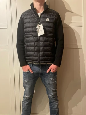 Svart cardigan från Moncler - Snygg svart dunväst från Moncler med quiltad front och stickade ärmar. Västen har hög krage, dragkedja och Moncler-logga på bröstet. Perfekt för kalla dagar och ger en clean, modern look. Materialet är en mix av dun och stickad textil.
