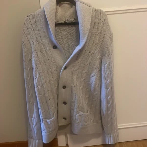 Ljusgrå kabelstickad kofta - Säljer en ljusgrå kabelstickad kofta med stora knappar och bred krage. Koftan har två fickor framtill och ribbade muddar vid ärmslut och nederkant. Perfekt för lager på lager och riktigt mysig att ha på sig. Super fint skick o inga defekter förutom att taggen ramlat av i tvätten. 