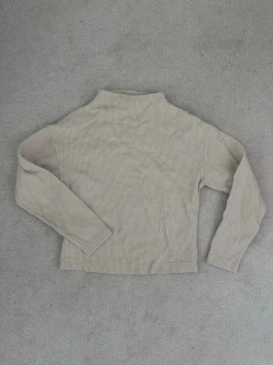 Beige ribbstickad polotröja - Säljer en beige ribbstickad polotröja med lång ärm. Tröjan har en hög hals och är tillverkad i ett mjukt, stretchigt material som sitter skönt på kroppen. Perfekt att styla med jeans eller kjol för en clean look.