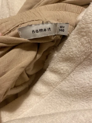Beiga linnebyxor från Name It - Säljer ett par beiga linnebyxor från Name It i storlek 140. Byxorna har rak passform, resår i midjan och är tillverkade i ett lätt och luftigt linnematerial. Perfekta för en avslappnad och stilren look.