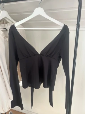 Svart tröja från Nelly - Snygg svart topp från Nelly med djup v-ringning och långa ärmar. Toppen har knytband i midjan för en bra passform och är tillverkad i ett mjukt, stretchigt material. Med rosett i ryggen💗🤩             Använd fåtal gånger!