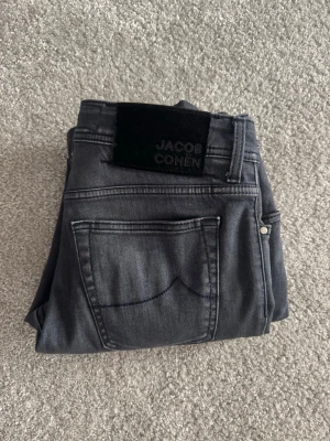 Grå jeans från Jacob Cohen - Snygga grå jeans från Jacob Cohen med klassisk femficksmodell och knappgylf. Jeansen har en diskret tvätt och svart patch med logga bak i midjan. Materialet är mjukt och stretchigt för skön passform. Perfekta till en avslappnad stil. Använda runt 5 gånger köpta nya.