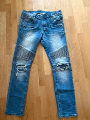 Balmain jeans - Storlek 33. Säljer ett par ljusblå bikerjeans från Balmain med slitningar och dragkedjor framtill. Jeansen har ribbade detaljer över knäna och trasiga partier på låren för en cool, edgy look. Klassisk femficksmodell och tillverkade i denim.