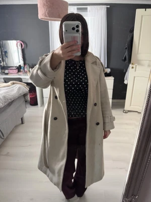 Beige lång kappa med dubbelknäppning - Stilren beige kappa i lång modell med dubbelknäppning framtill och breda slag. Kappan har markerade axlar och dekorativa remmar vid ärmsluten. Perfekt för dig som gillar en klassisk och tidlös look. Kappan är lite nopprig men inget man tänker på 