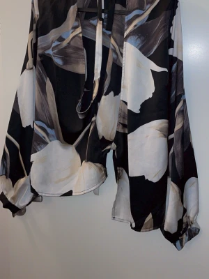 Blommig blus med knytband - Snygg blus med stora vita och beige blommor på svart botten. Blusen har långa, vida ärmar med resår vid handlederna och ett dekorativt knytband vid halsen. Tillverkad i ett tunt, flowy material som ger en avslappnad men trendig look.