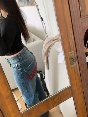 BDG Jeans - Säljer ett par blå BDG jeans med rak passform och slitna detaljer. På baksidan finns en stor röd brodyr med texten 'Bdg Jeans' över bakfickorna. Jeansen har klassisk femficksdesign och är gjorda i denim. Perfekta för en avslappnad streetwear-look. Storlek: 26W 32L/ storlek: 34/36. Original pris: 600-800kr.