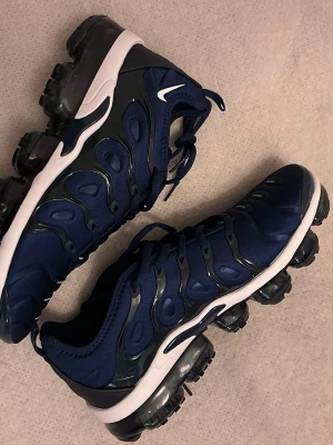 Nike Air Vapormax Plus marinblå sneakers - Nike Air Vapormax Plus sneakers i marinblått med svarta och vita detaljer. Ovandel i mesh och syntet med ikoniska vågformade paneler. Sulan har synliga luftbubblor för maxad komfort och en futuristisk look. Perfekta för dig som gillar streetstyle och sportig vibe.