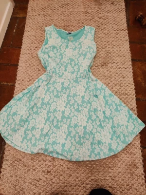 Turkos blommig klänning XXL  - Säljer en söt turkos klänning med vitt blommönster. Klänningen är ärmlös och har en rund halsringning samt utställd kjol. Perfekt för varma dagar och ger en fräsch look.