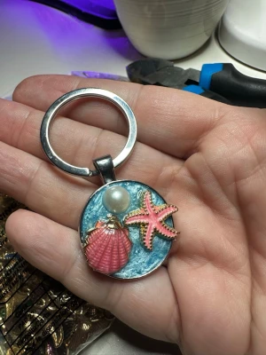 Nyckelring med snäcka och sjöstjärna - Cool nyckelring med havstema, dekorerad med en rosa snäcka, en rosa sjöstjärna och en pärla på en blå bakgrund. Metallring och detaljer i guld och silver. Perfekt för dig som gillar strandvibbar och unika accessoarer.