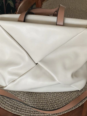 Handväska från Parfois - Härligt följsam väska i ljust konstläder med bruna detaljer. Storlek 35x24. Väskan är rymlig och har ett innerfack med dragkedja. Den är i bra skick men lite sliten på handtagen. Se sista bilden.