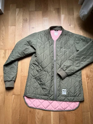 Olivgrön quiltad pufferjacka Kari Traa - Snygg olivgrön pufferjacka från Kari Traa  med quiltat mönster och rosa innerfoder. Jackan har dragkedja framtill, ribbade muddar vid ärmslut och längre bakstycke. Perfekt för våren / sommaren  och riktigt skön att bära.