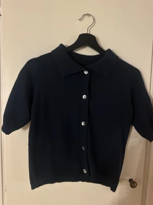 Mörkblå stickad kofta med kort ärm - Mörkblå stickad kofta med korta ärmar och klassisk krage. Koftan har knäppning framtill med silverfärgade knappar och en rak passform. Perfekt att styla över en t-shirt eller linne. Aldrig använd. 