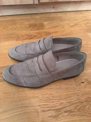 Grå mockaloafers - Säljer ett par stilrena loafers i grå mocka med klassisk pennyloafers-look. Skorna har rundad tå, låg klack och dekorativ söm längs ovandelen. Perfekta för dig som gillar en clean och tidlös stil.