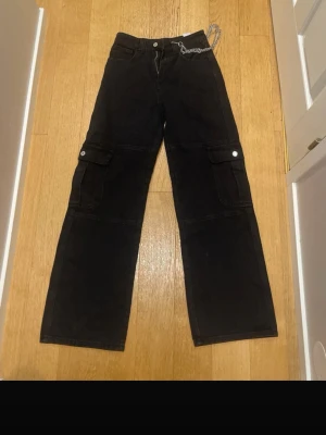 Svarta cargobyxor med kedja - Säljer ett par svarta cargobyxor med bred passform och stora fickor på sidorna. Byxorna har en snygg silverfärgad kedja i midjan som detalj. Tillverkade i ett kraftigt bomullstyg och har knapp- och dragkedjestängning. Perfekta för en streetwear-look.