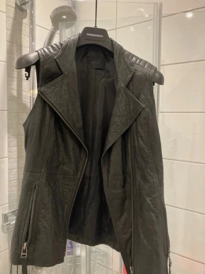 Svart skinnväst från Gestuz - Säljer en svart skinnväst från Gestuz med cool bikerstil. Västen har breda slag, dragkedjor framtill och detaljerade axelpartier. Perfekt för att lyfta din outfit med en edgy touch. Materialet är mjukt och följsamt konstläder.