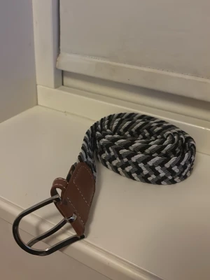 Randigt flätat bälte med läderdetalj - Snyggt flätat bälte i svart, vitt och grått med bruna läderdetaljer vid spännet. Bältet har ett klassiskt metallspänne och passar perfekt för att ge din outfit en extra touch. Flexibel och justerbar längd tack vare det elastiska materialet.