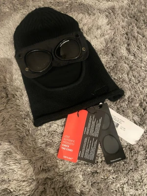 Svart goggle-mössa från C.P. Company - Säljer en svart stickad goggle-mössa från C.P. Company. Mössan har inbyggda glasögon med logga . Tillverkad i mjukt, ribbstickat material som ger en cool och unik look. Perfekt för dig som vill sticka ut med streetwear-stil.