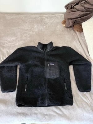 Svart fleecejacka från Patagonia - Mysig svart fleecejacka från Patagonia med hel dragkedja, hög krage och tre fickor med dragkedja. Bröstfickan har ett snyggt tygparti och logga. Perfekt för lager-på-lager och riktigt skön att ha på sig under kyliga dagar. Storlek M men liten i storleken så passar även S!