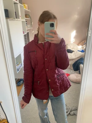 Vinröd quiltad jacka med dragkedjor - Snygg vinröd quiltad jacka med ståkrage och flera dragkedjefickor framtill. Jackan har knappar och dragkedja som stängning samt en figurnära passform. Perfekt för kyligare dagar och enkel att matcha med jeans.