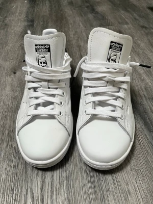 Adidas Stan Smith vita sneakers - Klassiska Adidas Stan Smith sneakers i vitt skinn med svart detalj på hälen och ikonisk logga på plösen. Skorna har snörning och en stilren, tidlös design som passar till det mesta. Perfekta för dig som gillar en clean och sportig look. Nyskick, aldrig använda. Nypris:1299. Mitt pris:550. Pris går att diskutera vid smidig affär. 