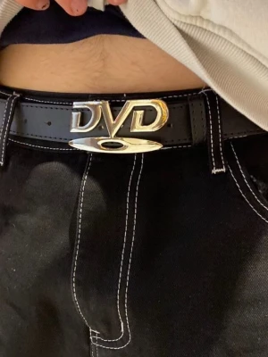 Svart Y2K bälte med DVD-spänne - Säljer ett svart bälte med ett stort, blankt metallspänne där det står DVD. Sparsamt använd. Längd 105 cm