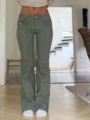 Gröna bootcut jeans med fickdetaljer - Snygga gröna bootcut jeans med låg midja och dekorativa fickor framtill. Jeansen har vida ben och dubbla knappar i linningen för en trendig look. Perfekta för dig som gillar 00-talsstil och vill sticka ut.