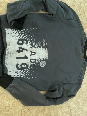 Mörkgrå sweatshirt från Stone Island - Cool mörkgrå sweatshirt från Stone Island med stor grafisk print och texten 6419 på framsidan. Tröjan har rund halsringning, svarta muddar vid ärmslut och nederkant samt en avslappnad passform. Perfekt för dig som gillar streetwear och unika detaljer.
