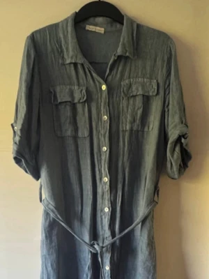 Blå långklänning i linne med knappar - Snygg grå långklänning i linne med knappar hela vägen framtill, två bröstfickor och knytskärp i midjan. Klänningen har uppvikta ärmar och avslappnad passform, perfekt för en stilren look. Hade på mig en gång, dubbelt storlek L/XL. Klänningen kostade 1099kr, nu 500kr, ej prutbar.