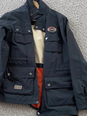 Blå vindjacka från Morris - Snygg blå vindjacka från Morris med flera praktiska fickor framtill och coola patchdetaljer. Insidan är beige och orange för en schysst kontrast. Jackan har tryckknappar och dragkedja, samt justerbar nederkant. Perfekt för dig som vill ha en stilren och funktionell jacka.