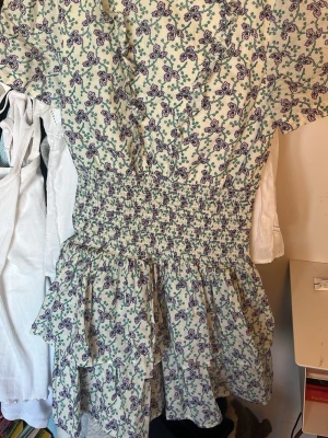 Blommig klänning  - Supersöt kort klänning från Les Coyotes de Paris med lila blommönster på ljus botten. Klänningen har volanger framtill, smock i midjan och en  kjol med volangkant. Säljs inte längre❣️Klänningen har en liten fläck på bröstet/magen men det är inget som man ser! Fråga gjärna så skickar jag bild! Står ingen storlek, men skulle säga xxs/xs