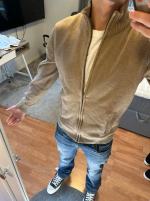 Beige stickad ziptröja med hög krage - Säljer en beige stickad ziptröja med hög ribbad krage och dragkedja framtill. Tröjan har två fickor på sidorna och ribbade muddar vid ärmslut och nederkant. Perfekt för lager på lager och enkel att matcha med jeans.