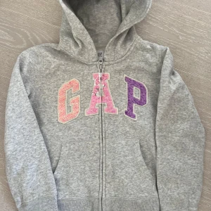 Grå hoodie från GAP Kids - Säljer en ljusgrå/vit hoodie från GAP Kids med dragkedja och färgglada paljettbokstäver på bröstet. Perfekt för en avslappnad stil. Den har en praktisk huva och långa ärmar.