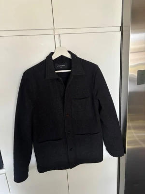 Mörkgrå overshirt från A Day's March - Stilren mörkgrå overshirt från A Day's March i ullmix. Klassisk krage, tre framfickor och knäppning med svarta knappar. Perfekt lager-på-lager-plagg med rak passform och diskret design som funkar till det mesta.