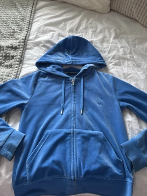 Blå velour hoodie från Juicy Couture - Säljer en blå velour hoodie från Juicy Couture med dragkedja framtill, fickor och justerbar huva med snören. Klassisk JC-logga broderad på bröstet och ribbade muddar vid ärmslut och nederkant. 